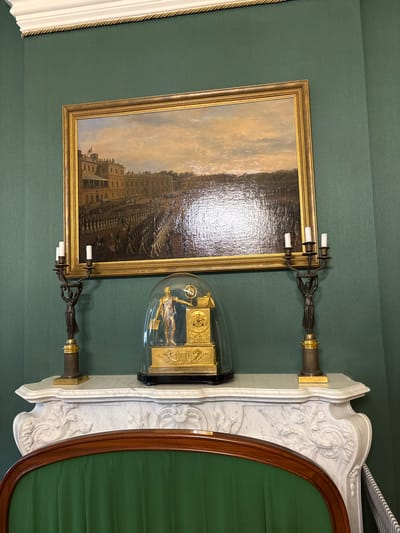 A gilded frame painting of a parade before a grand building hangs on a dark green wall. Below the painting, a white marble mantelpiece features a gold clock under a glass dome. The clock depicts a uniformed figure with a globe. To the left and right of the clock are ornate candelabras with three candle holders each. The scene is located in the Knights' Hall of the Gatchina Palace, Gatchina, Russia. A green upholstered sofa is partially visible in the foreground.