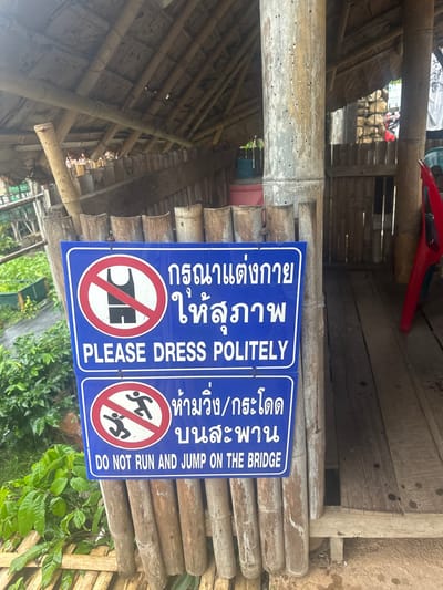 The image displays a blue informational sign, centrally located and affixed to a weathered vertical bamboo pole, within a rustic structure in Ban Paem Bok, Thailand. The sign is divided into two sections. The upper section features a red circular prohibition symbol over a white outline of a sleeveless top and shorts, accompanied by Thai text and the English phrase "PLEASE DRESS POLITELY." The lower section presents a red circular prohibition symbol over white outlines of two running/jumping figures, with Thai text and the English phrase "DO NOT RUN AND JUMP ON THE BRIDGE."
The sign and bamboo pole are situated under a sloped, thatched roof supported by a bamboo framework. To the immediate left of the sign, a low fence composed of vertical bamboo segments runs horizontally. Beyond this fence, green leafy vegetation, possibly coffee plants, is visible. To the right of the sign, a wooden plank floor extends into the background, where a partially visible red plastic chair and a blue container are discernible. The main bamboo pole exhibits numerous small, dark circular marks, indicative of wear or insect activity. No individuals are present in the scene.
