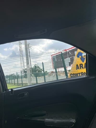 The image depicts a view through the rear window of a vehicle, looking out towards a street in Wupa, Nigeria. The foreground is dominated by the interior of the car, including the dark-colored door panel with a black handle and window controls. Outside the window, a chain-link fence runs horizontally across the frame, with a tall, grey pole to the left, likely a utility pole, possibly with a cell tower in the background. Behind the fence, a large yellow billboard with blue and black lettering is visible, with the words "ARA CONTR" partially legible. The billboard appears to be in front of a larger, dark-colored structure or sign. The sky is partly cloudy.