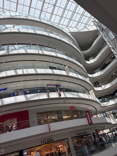 Here is a description of the image:
The image depicts the interior of a modern shopping mall, likely taken from a high vantage point looking downwards. The architecture features multiple levels with curved, white balustrades. Natural light streams in through a large grid-patterned window structure on the left. Several retail storefronts are visible, indicated by brand name signs: "HUGO," "MAISON DE PARFUM," "ALVIERO MARTINI 1 CLASSE," "INTERSPORT," "SCOTCH & SODA," "BEE KIDS," and "EPIA IDEA." A partial sign reads "SPORT" on the bottom-left.
Based on the storefront signage, the shopping mall is likely located in the city of Tirana, Albania.