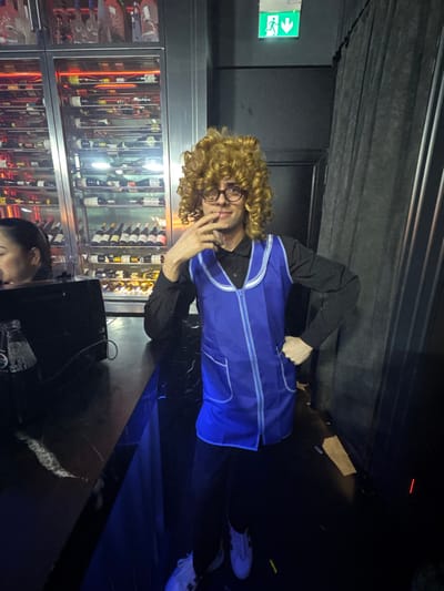 The scene is set indoors, likely within a bar or lounge in Dubai, United Arab Emirates. A male individual stands centrally, facing the camera. He wears a light brown, shoulder-length curly wig, round eyeglasses, a black long-sleeved shirt, dark trousers, and white sneakers with black accents. Over his shirt, he sports a vibrant blue sleeveless apron or tunic with white piping, a front zipper, and two lower pockets. His right hand is posed with fingers touching his chin, and his left hand rests on his hip. He displays a slight smile. To the left, a female individual with dark hair is partially visible from the chest up, looking downwards at a dark, reflective bar counter. On the counter, a dark electronic device and a clear bottle are present. Behind the primary subject, a large, illuminated glass display cabinet filled with numerous bottles of wine and spirits arranged horizontally is prominent. To the right, a dark, heavy, vertically pleated curtain is visible. A green emergency exit sign, featuring a white running man pictogram and a downward arrow, is mounted on the dark wall above the main subject's head. The environment is dimly lit, with specific illumination from the beverage display and the exit sign.