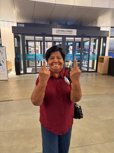 A medium shot captures a woman standing outside "Los Balcones Restaurants y Cafés," a business located in the city of Alcorcón, Spain. The woman, with dark hair and a smiling expression, is wearing a maroon button-up shirt and blue jeans. She is holding up both of her middle fingers towards the camera. A black handbag hangs from her right shoulder. The entrance to "Los Balcones Restaurants y Cafés" is visible behind her, with glass doors and signage. The floor is tiled, and a trash receptacle and a wooden cabinet are present to the right of the entrance.