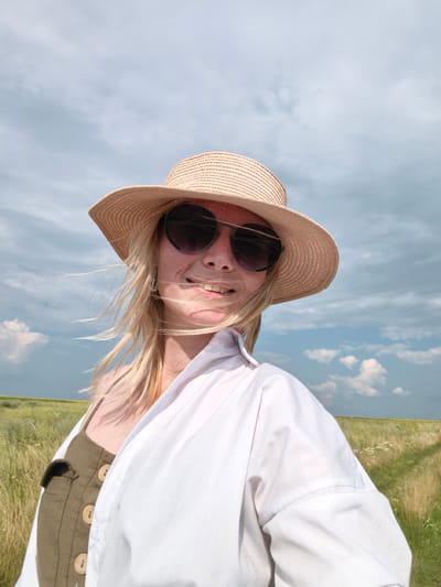 The image is a portrait of a woman wearing a straw hat and sunglasses, taken in a grassy field with a cloudy sky. The woman has blonde hair and is smiling. She is wearing a white button-up shirt over a green tank top with wooden buttons. The background shows a wide expanse of tall grass and a distant horizon. The location is identified as Kuzminovka, Russia.