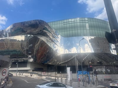 The image captures the exterior of a modern building in Birmingham, United Kingdom, characterized by its highly reflective, undulating metallic facade. The building appears to be part of a larger complex, with a glass-paneled upper section visible above the metallic curves. Reflected in the metallic surface are distorted images of surrounding architecture and the sky.
In the foreground, a paved street or sidewalk leads towards the building. There are several bollards lining the street and a traffic light is visible. A white Tesla Model 3 is parked on the right side of the frame, and another vehicle, possibly a dark sedan, is seen in the background on the left. A sign with the text "GRAND CENTRAL P" indicates a parking area.
Two individuals are walking on the sidewalk on the left side of the image. One person, wearing a black and white checkered dress, is walking away from the camera, while the person next to them is partially obscured.
Below the reflective facade, a section of the building features windows and signage. One prominent sign reads "WETHERSPOON," indicating a pub or restaurant. Further signage suggests "Welcome to Birmingham New Street Station" and provides directions to a car park and drop-off points.
The environment appears to be an urban setting, with a bright sky and scattered clouds suggesting daytime. The architecture is contemporary, with the metallic structure forming a striking visual element.