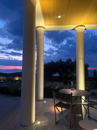 In the Municipal Unit of Kranidi, Greece, at twilight, an outdoor seating area is depicted. Large, cream-colored pillars support a wooden ceiling with recessed lighting. In the foreground, a round, dark gray table is set with a small, modern portable lamp. Two foldable director's chairs with dark gray canvas seats and backs and wooden frames are positioned at the table. The floor is paved with light-colored marble tiles. Beyond the seating area, the landscape features sparse, dry grasses and white flowers. In the background, a dark silhouette of an olive tree stands against a sky transitioning from deep blue to hues of purple and pink as the sun sets over distant mountains and a body of water. Several ground lights are visible on the patio, illuminating the pillars and the immediate surroundings.