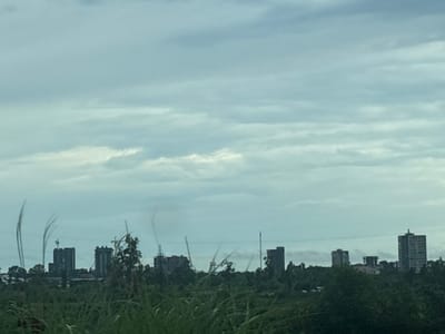 The image depicts a cloudy sky over a cityscape in Kobuleti, Georgia. In the foreground, tall green grass and trees obscure the lower portions of the scene. Silhouetted against the sky are several multi-story buildings, some appearing to be under construction with cranes visible. Thin, wispy lines, possibly indicative of smoke or atmospheric phenomena, are present in the sky above the buildings. The overall impression is one of a developing urban area under an overcast sky.