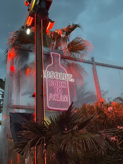 The image shows a neon sign for Absolut Vodka in Alimos, Greece. The sign, shaped like a vodka bottle with the text "ABSOLUT. BORN TO DREAM" is mounted on a structure behind palm trees. Additional lighting fixtures shine on the trees and sign. The background is an overcast sky.