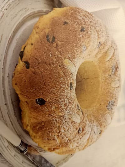 A bundt cake is placed on a white, distressed wooden platter. The cake is golden brown with visible raisins and almond slivers embedded in its surface. A light dusting of powdered sugar covers the cake. The platter has a slightly raised rim and appears to be resting on a white textured fabric, possibly a tablecloth or napkin. To the left of the platter, a knife with a white handle and a serrated blade rests partially on the platter and partially on the table surface. The background is a soft, white fabric. There is no discernible location in the city of Rumaithiya, Kuwait, or any people present in the image.