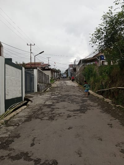 This image depicts an uphill residential street in Melakudikadu, India, under an overcast sky. The dark gray asphalt road surface is visibly worn and patched. On the left, a white perimeter wall with a distinct wave-patterned texture borders the street, featuring a dark gray gate. Beyond this, various residential buildings with different rooflines are visible. Multiple utility poles support a complex network of overhead electrical and communication wires, with a streetlamp attached to one pole. A small red and white banner is visible further up the street on the left. On the right side, an elevated embankment, partially vegetated, supports more residential structures; laundry is seen hanging from one building. A child, wearing a green patterned shirt and blue shorts, is running away from the viewer up the street, on the right side of the road. Further up the street, a dark-colored vehicle is present, and an adult in red clothing is walking on the right side.