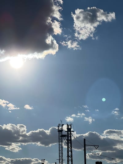 The image captures a sky scene in Újezd u Brna, Czechia. Silhouetted against a bright sky filled with scattered clouds, several structures are visible. In the foreground, two tall, dark, lattice-like towers stand, one on the left and another partially visible on the right. Power lines crisscross the frame between these structures. The sky is a mix of bright blue and areas obscured by clouds. A prominent, bright sun is visible in the upper left quadrant, with a lens flare creating a luminous effect and a faint contrail from an aircraft. In the upper right quadrant, a distinct, teal-colored circular artifact is present, possibly a reflection or lens anomaly. The clouds are varied in density, with some appearing dark and heavy, while others are lighter and wispy, catching the sunlight. The overall impression is of an outdoor scene, possibly taken from an elevated position, with a focus on the sky and man-made infrastructure.
