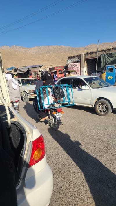 The image shows a street scene in Kuchlak, Pakistan, likely a marketplace. Several vehicles are visible, including white cars and colorful auto-rickshaws, some heavily loaded with goods and passengers. A motorcycle has a blue frame attached for carrying cargo, which includes black bags and white cloth. People are dressed in traditional clothing. The background features a clear blue sky and a landscape with bare mountains. The road is paved.