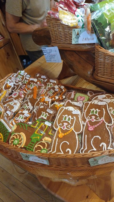 The image shows a close-up view of a basket filled with intricately decorated gingerbread cookies, predominantly in the shapes of animals like dogs and cats, as well as a carriage. The cookies are individually wrapped in clear plastic and have prices marked on them, such as "3.00" and "4.00". There are also some colorful candies in cellophane wrappers visible in a basket behind the gingerbread. A label on the basket reads "LATVIJA" with a small illustration of a fairy. In the background, a person wearing a light brown t-shirt is partially visible, with their arms blurred as they appear to be engaged in an activity. The setting appears to be a market or shop, with wooden surfaces and a rustic ambiance. While the specific location within Tērvete, Latvia is not explicitly stated, the "LATVIJA" label suggests a connection to the country.