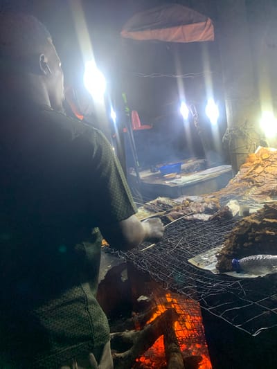The image depicts an outdoor food stall at night, likely in Port Harcourt, Nigeria, with several bright lights illuminating the scene. A person, seen from the back and side, is wearing a dark green, patterned short-sleeved shirt. They appear to be tending to a grill laden with various types of meat, some of which are skewered. A fire burns beneath the grill, casting a warm glow. Smoke rises from the food, indicating it is actively being cooked. In the background, more food is visible on a counter, along with a blue plastic bowl and a water bottle. The environment suggests a street-side food vendor operation, common in many urban areas of Nigeria.