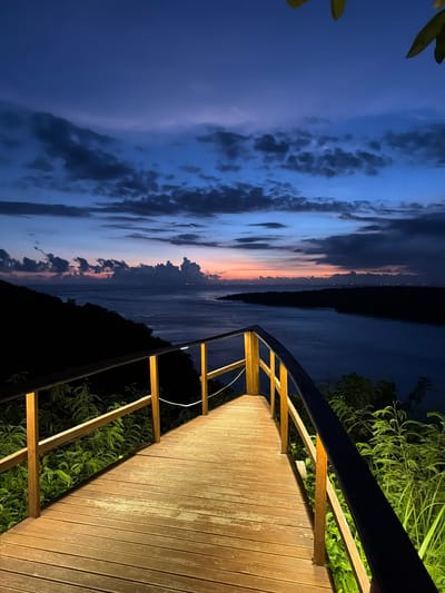 The image depicts a boardwalk with a railing leading towards a view of a body of water and a colorful twilight sky. The boardwalk is made of wooden planks and is illuminated by warm lighting. Vegetation is visible on both sides of the boardwalk, suggesting a natural environment. The sky displays a gradient of colors, transitioning from dark blue to shades of pink and orange near the horizon, indicating either a sunset or sunrise. The water appears calm. The scene is likely located in Sakti, Indonesia, given the location data.