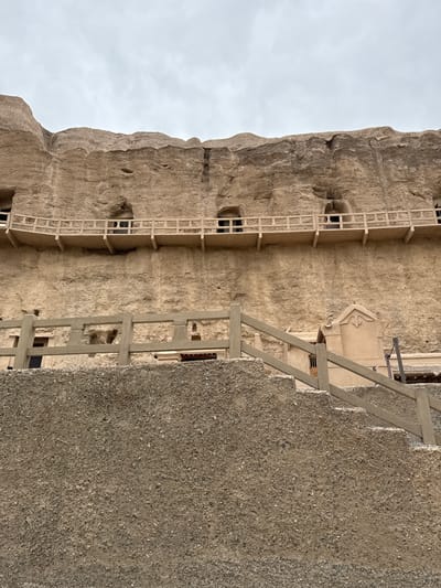 The image shows a rock-cut architecture, possibly a series of cave temples or dwellings. It is situated in Guazhou County, China. A stone structure with scaffolding is visible. The architecture appears ancient, with elements such as the carved facades and the weathered texture of the rock. The presence of ladders and platforms suggests ongoing preservation and restoration efforts. The sky is overcast. A wall of some kind extends from the right side of the picture.
