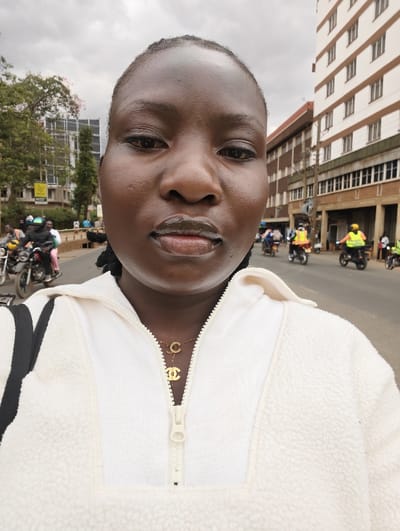 A medium close-up, front-facing selfie captures a woman in an urban setting under an overcast sky. The woman, who has dark skin, is centered in the frame, looking directly at the camera with a neutral expression. Her hair is pulled back, and her facial features include dark eyes and full lips, which appear glossy and have a lighter substance visible on the lower lip.

She is wearing a cream or off-white fleece jacket with a half-zip neckline. A golden necklace rests on her chest, adorned with two charms: a single capital 'C' and the interlocking 'CC' logo, indicative of the Chanel brand.

The background reveals a bustling street scene with several motorcycles and people. On the left side, vehicles and pedestrians are visible, along with trees, one of which has faint pink blossoms. Behind the trees, tall buildings rise, including a modern-looking structure with reflective surfaces. A partially visible yellow sign with black text reads "NCCE".

On the right side of the background, more motorcycles are seen, with several riders wearing distinctive bright yellow safety vests. Further back, a row of buildings lines the street, featuring both older and more modern architectural styles, including a prominent multi-story building with numerous windows. The sky overhead is uniformly cloudy, suggesting an overcast day rather than bright sunshine.