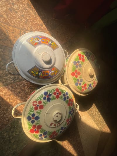 Three lidded, enamelware food containers are positioned on a tiled floor in Isiamigbo, Nigeria. The largest container at the top features a white lid with a colorful, segmented abstract pattern incorporating stylized faces. Two smaller containers below it have white lids decorated with a repeating floral motif of red and blue flowers with green leaves. The lid of the bottom container explicitly shows the letters "FAO" in red. All containers have a central lid handle and two side handles on the pot body. Strong, directional sunlight illuminates portions of the scene, creating sharp shadows on the speckled, patterned floor tiles. No individuals or active interactions are present.