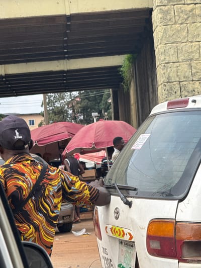The image captures a bustling street scene under an overpass in Orji, Nigeria. In the foreground, the back of a white Toyota car is visible, with a sticker on its rear window and a license plate indicating "ENUGU" and "GOVERNMENT STATE." A person wearing a colorful, wavy-patterned shirt and a black baseball cap with the "POLO" logo is in the mid-ground, facing away from the camera. Behind them, two red umbrellas are set up, likely for market stalls, with people interacting and standing around. The overpass structure is prominent, made of concrete and steel beams, with brickwork on one side. The environment suggests a typical urban setting with ongoing street activity and possibly a local market.