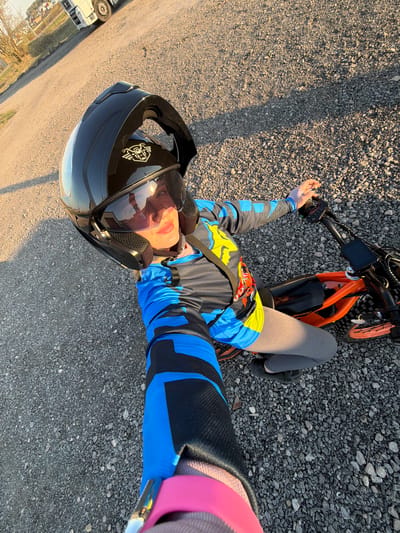 The image shows a person lying on a gravel road, wearing a black motorcycle helmet, a blue and black long-sleeved shirt, gray leggings, and black shoes. The person is next to an orange electric bike. The photo appears to be a selfie taken with the person's arm extended. The background shows a road leading to what seems to be a truck and some buildings. The location is indicated as Азьмушкино, Russia.