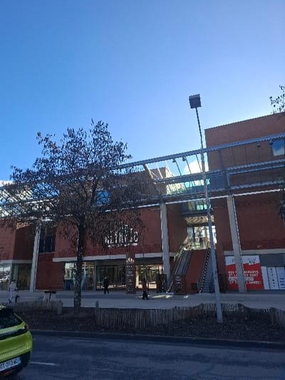 The image displays the exterior of the Centre Commercial Grand'Place in Saint-Martin-d'Hères, France, under a clear, bright blue sky. The multi-story building features a facade of red-brown brick, large glass windows, and a prominent steel and glass canopy structure. The name "GRAND PLACE" is visible in white lettering above the central entrance.
In the foreground, a dark asphalt surface, likely a road or parking area, runs horizontally. The rear of a lime-green passenger car, with a partially visible license plate "GT-263-WZ," is positioned on the far left. A low wooden picket fence separates the asphalt from a pedestrian area with dark ground cover, possibly mulch, and a bare-branched deciduous tree centrally located on the left side of the frame.
Several individuals are present on the paved pedestrian walkway in front of the building. One person in light-colored clothing walks left on the far left, while two others approach the main entrance near the center. On the right side of the building's facade, an outdoor escalator system leads to an upper level. Below the escalator, a red commercial sign is visible, partially obscured, promoting restaurants open "7J/7 ET TOUTE L'ANNEE!" A tall, dark lamppost stands vertically in the right-center of the image, extending into the sky. The scene is illuminated by strong daylight, indicating a daytime setting.