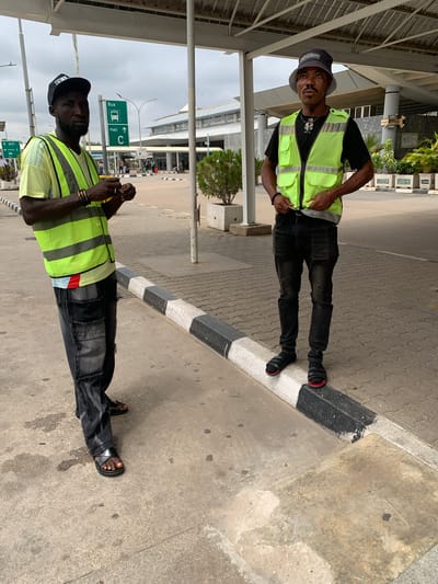 Two men wearing neon yellow safety vests stand on a paved surface next to a road, possibly in Gwaska, Nigeria. The image appears to be taken during the daytime under an overcast sky.
The man on the left wears a dark bucket hat with the word "AVEC" visible, a black t-shirt, dark pants, and black sandals with red accents. He has a light mustache and is looking towards the camera with a slight smile, his hands resting on his vest pockets. He wears red and black bracelets on his left wrist.
The man on the right wears a black baseball cap with "ELI" or "FFL" visible, a black t-shirt, dark pants with textured panels, and black sandals. He has a beard and is looking slightly downwards or forward, holding his hands together in front of him. He wears dark bracelets or rings on his wrists.
They are positioned near a black and white striped curb that separates the paved area from a concrete section. In the background, there is an overhead structure, various buildings, and some greenery. Prominently visible are green traffic signs indicating directions: one reads "Bus" with a bus icon and "Hall" with an upward arrow, and "C" also with an upward arrow. Another smaller sign below it shows "Exit" and "C" with arrows, suggesting a transportation hub like an airport. In the far background, stacked items with the word "HERBAN" are faintly visible.
