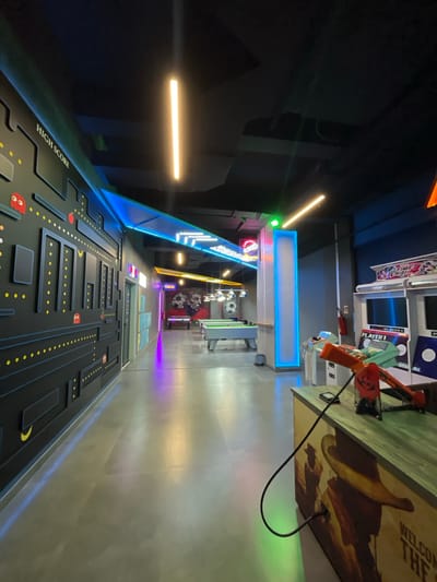 The image captures the interior of an arcade or entertainment facility, possibly located in Mechouar Kasbah, Morocco. The perspective is from a low angle, creating a sense of depth and emphasizing the arcade's layout.

The foreground features a black wall with a stylized design resembling a Pac-Man maze, complete with the "HIGH SCORE" label printed on it. Recessed black rectangles, yellow dot motifs, and stylized red ghost figures decorate the surface.

Moving toward the center, the image reveals various arcade game cabinets. A pair of orange toy guns are visible atop one of the arcade machines. The arcade cabinets display game titles.

The ceiling has blue and white neon lighting fixtures, which illuminate the corridor. The walls appear to be painted in light, metallic shades, reflecting the arcade's lighting.

The floor is made of a smooth, light-colored material, likely concrete or tile. Further down the corridor, there are decorative elements, including signage and potentially more arcade games.

The environment appears to be a modern and well-lit arcade or entertainment center, with a focus on interactive gaming experiences and a retro arcade aesthetic.