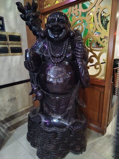 A dark-colored statue of a smiling Buddha stands indoors in Chu Văn An Ward, Vietnam. The statue is adorned with a necklace and holds objects in its hands. It has a large belly and is mounted on a tiered base. Behind the statue are walls of light neutral color with electrical outlets, a dark-colored frame surrounding decorative gold-colored panels with green and blue background. A brown door frame is visible to the right. The floor is covered with light-colored tiles.