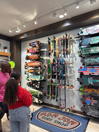 The photograph shows the interior of a surf shop in Lake Buena Vista, Florida, United States, displaying various skateboards mounted on the wall. The skateboards vary in design, size, and color, arranged in multiple rows. A "Ron Jon Surf Shop" logo is visible on the skateboards and a blue rug.
In the foreground, a group of people is visible, including a person wearing a pink shirt, a person wearing a red hoodie and jeans, and a person with a shaved head wearing a lime green hat. The person in the red hoodie appears to be bending over or looking at something near the bottom row of skateboards. The scene suggests a retail environment where customers are browsing merchandise.