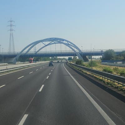 This is a frontal, eye-level photograph taken from a vehicle moving on a highway. The road surface is asphalt with visible white lane markings. A metal guardrail runs along the right side of the road. In the background, a large, modern bridge with twin arched spans, painted in light blue and white, crosses the highway. Below this bridge, a separate, straight blue bridge is visible. To the left of the highway, a tall, lattice-style electrical pylon stands against a clear, pale blue sky. A red truck is positioned on the road beneath the arched bridge, and several cars are also visible on the highway, heading in the same direction as the photographer. The surrounding environment appears to be rural or semi-rural, with sparse vegetation and trees visible beyond the highway. The overall impression is one of a bright, sunny day on a major transportation route. The location is identified as being in the municipality of Montirone, Italy.