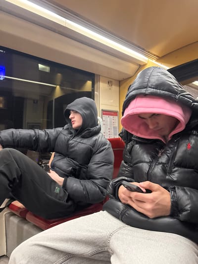 Two individuals are visible inside what appears to be a train or bus. One person is seated, wearing a black puffer jacket with a pink hood underneath. They appear to be sleeping. Another individual beside them is also wearing a black puffer jacket and grey sweatpants. Another person is seated across the aisle in a black jacket and sweatpants, facing forward. The interior consists of beige walls, a window, and informational signage.
