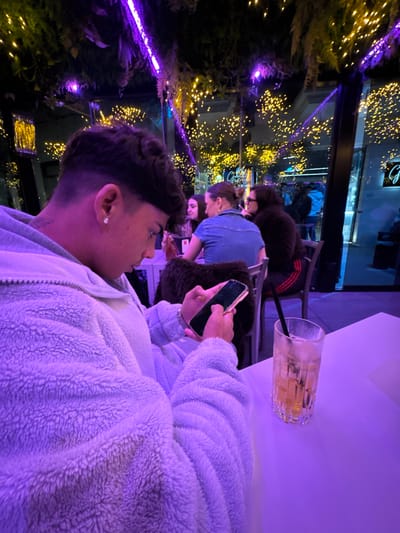 This image depicts an indoor scene within a bar or restaurant in Stornara, Italy, illuminated primarily by purple and yellow artificial lights at night.

In the foreground, a young individual, presumed male, with dark, styled hair and an earring in his left ear, is seated at a white table. He wears a light-colored, possibly sherpa or fleece, zip-up jacket and is actively engaging with a dark-screened smartphone held in both hands. A tattoo is visible on his left arm. To his right, on the white table, sits a tall, clear glass containing an amber-colored beverage with ice cubes and a black straw.

In the middle ground, several other individuals are seated at tables. One person, wearing a denim top, is positioned with their back mostly to the camera, appearing to interact with another individual seated opposite them, who wears a dark top. Additional patrons are dimly visible further in the background.

The environment features an overhead canopy decorated with artificial green foliage, interspersed with numerous small yellow string lights and prominent purple neon light strips. Large glass windows or panels form the background, revealing reflections of the interior lighting and a blurred view of an exterior street or area. An illuminated sign with the letter "G" is discernible through the glass, suggesting another establishment or business nearby. The overall lighting casts a dominant purple hue across the foreground and midground areas.