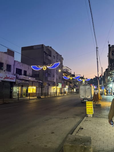 This image captures a street scene in Downtown Amman, Jordan, at twilight or dawn, indicated by the dark blue sky transitioning to an orange hue near the horizon. The street, which appears damp in sections, runs through the center of the frame, bordered by multi-story, aged buildings on both sides. Overhead, multiple blue and yellow decorative light installations, shaped like crescent moons with a central orb, are strung across the street.
On the left, several ground-floor shops are visible; one with a sign reading "الشاطر" (Al-Shater) has its metal shutter mostly closed, while another nearby shop displays illuminated merchandise through its grates. Further down the street on the right, a white enclosed delivery truck is parked, facing away from the camera. Adjacent to the truck, a yellow advertising sign points right, displaying Arabic text and a Jordanian phone number (0776347792). Garments are hung on a display stand on the sidewalk next to the truck.
The foreground on the right features a paved sidewalk with a concrete block, and the lower right corner shows the torso and arm of an individual carrying a plastic bottle, walking away from the camera. Overhead power lines crisscross the upper portion of the frame. Streetlights are illuminated, casting warm light on the immediate surroundings and the street surface.