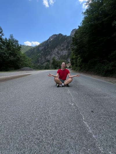 A young woman wearing a red t-shirt and shorts sits cross-legged on an asphalt road, performing a meditation pose. She has glasses on and her hands are resting on her knees with her thumbs and index fingers touching. The road appears to be in a mountainous, forested area, possibly a remote part of Crna Poda, Montenegro, based on the surrounding natural landscape. Tall, rugged mountains rise in the background, covered in trees, and a clear blue sky with a few scattered clouds is visible. The road is empty of traffic and has a white line marking one side. The overall scene conveys a sense of peace and solitude amidst nature.