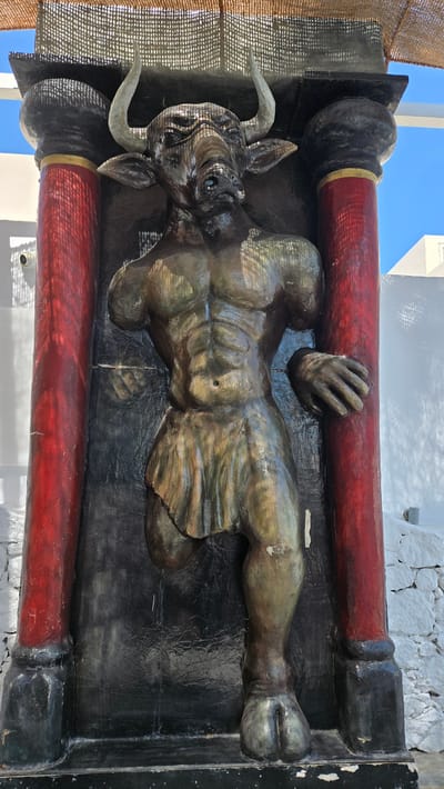A statue of the mythological creature the Minotaur, with the body of a man and the head of a bull, stands on a pedestal between two large red columns. The statue is made of a dark, weathered material and depicts the Minotaur with muscular arms, a skirt-like garment around its waist, and hooved feet. Its head is turned slightly to the left, and its eyes are partially closed. The statue is located in the city of Agía Marína, Greece, and appears to be an outdoor decoration. The background shows a white wall with some texture and shadows cast by an overhead canopy. The sky is visible at the top of the image.