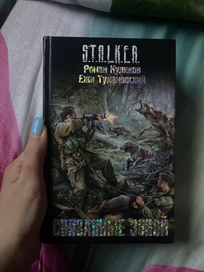 A book with a dark, illustrated cover is shown, held by a hand with light blue, glittery nail polish, resting on a patterned surface of pink, white, and teal fabric. The image is oriented sideways, making the book appear on its side.

The cover art depicts a grim and intense scene within a desolate, overgrown forest. In the foreground, a man in tactical gear, including a vest and backpack, lies prone, aiming a rifle from which a muzzle flash or light emanates. Behind him, to the right, a second man is visible, appearing to be bound or entangled with rope, looking downwards in a distressed or injured state.

Several grotesque, mutated wolf-like creatures with sharp teeth and decaying features emerge from the dark foliage, seemingly attacking or approaching the men. A large creature with bared fangs is prominent in the upper center, while another smaller one is near the foreground character. The overall atmosphere conveyed is one of danger, struggle, and survival in a hostile, post-apocalyptic or wild environment.

Visible text on the book cover includes the main title "STALKER," rendered vertically in a holographic, reflective font on the left side. Below this, the horizontal names "РОМАН КУЛИКОВ" (Roman Kulikov) and "ЕЛЕНА ТУМАНОВСКАЯ" (Elena Tumanovskaya) are visible, likely identifying the authors. On the right side of the cover, another vertical block of holographic text is present, but largely illegible due to light reflections and image quality.

The general location context for this media file is Verkhnevesyoloye, Russia.