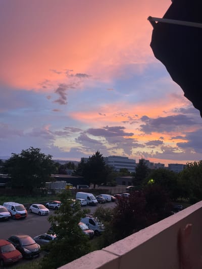 This vertical, full-shot photograph captures a sunset over an urban area, likely Vénissieux, France, as suggested by the presence of a row of parked cars, some with company logos. The sky is a vibrant display of pink, orange, and purple hues, transitioning into darker blues and grays as it reaches the horizon. Clouds are scattered across the sky, illuminated by the setting sun, creating dramatic shadows and highlights.

In the foreground, a balcony railing, painted in a pale pinkish hue, occupies the bottom right portion of the frame. Below the railing, several cars are parked on a tarmac lot. Among them are white vans, a red hatchback, a black hatchback, and a silver sedan. Some of the vans appear to be commercial vehicles, with one displaying the logo "ASELEC".

Beyond the parking lot, trees of varying sizes and densities are visible, some with dark green foliage, others with a reddish-purple hue. Behind the trees, a cityscape unfolds, with several modern buildings, including a large, multi-story structure with a reflective facade. The buildings are silhouetted against the colorful sky, with some windows appearing illuminated. In the distance, faint outlines of hills can be seen against the horizon. The overall scene suggests a twilight hour, with artificial lights beginning to appear in some of the buildings, contrasting with the natural light of the sunset.