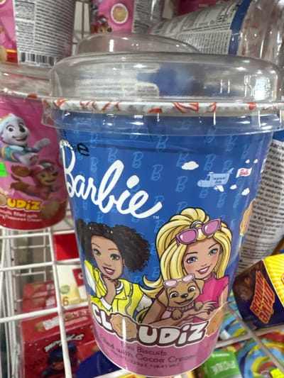 This is a close-up, slightly angled shot of several food items displayed on a store shelf in Alakhadzi, Abkhazia. The primary focus is on a cylindrical container with a clear plastic lid, adorned with colorful graphics of Barbie dolls and a puppy. The container's blue background is decorated with repeating "B" logos and small images of airplanes and clouds. The text "Barbie" is prominently displayed in white script. Adjacent to this, another similar container is partially visible, featuring graphics of characters that appear to be from Paw Patrol, with text that includes "JUDIZ" and possibly "PICTALKIE". Other packaged snacks with bright colors are visible on the shelf behind and around these items, suggesting a supermarket or convenience store setting. The lighting is bright, indicating an indoor setting, possibly during daylight hours. There are no discernible weather conditions or specific time of day cues beyond the indoor lighting. The overall impression is of a retail environment with a variety of children's-themed snack products.
