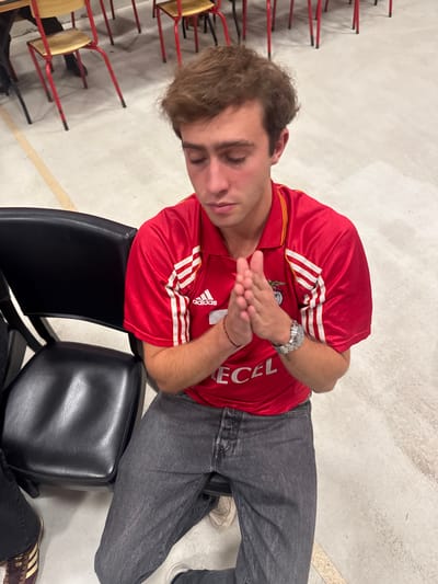 The image depicts an indoor scene in what appears to be a public or institutional building, such as a multi-purpose hall or gymnasium, within the city of Décines-Charpieu, France. The exact location within the city is not discernable from the visual evidence.
The central subject is a young adult male, seated with his head bowed and eyes closed. His hands are clasped together in front of his chest, fingers interlocked. He is wearing a red short-sleeved collared shirt with white shoulder stripes, an Adidas logo, a crest resembling the SL Benfica eagle, and the text "ECEL" printed on the front. A silver-toned watch with a metallic band is on his left wrist, and a thin dark bracelet is on his right wrist. He is dressed in dark grey jeans with a visible button fly. Light-colored sneakers are visible on his feet.
To the left of the central figure, a black upholstered chair with a backrest is partially visible. In the background, numerous light-colored wooden chairs with red metal frames are arranged on a light grey floor, which features a single yellow line marking. The lower left quadrant shows the foot of another individual, clad in dark trousers and brown sneakers with gold stripes. The environment is uniformly lit, indicating artificial indoor lighting.