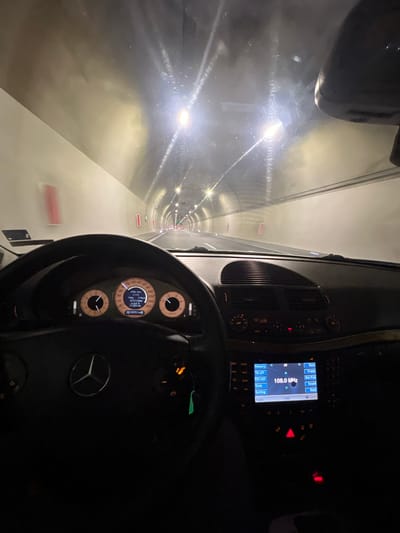 The image depicts the interior of a Mercedes-Benz car driving through the Jeleznitsa Tunnel in Jeleznitsa, Bulgaria. The tunnel is illuminated by bright overhead lights, and the car's dashboard is visible, showing the steering wheel with the Mercedes logo, the instrument cluster, and the infotainment system displaying the radio frequency. The car is moving through the tunnel.