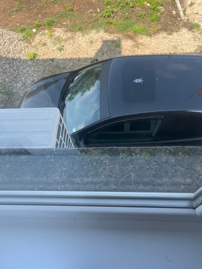 The image provides an overhead view from a window, looking down at a dark-colored car parked on a gravel and dirt surface. The scene is illuminated by strong daylight, indicating a sunny day.
In the foreground, a white window frame and a dark, textured sill or wall section are visible. This dark surface shows signs of weathering, with patches of green and yellowish moss or lichen. Partially visible near the window opening is a white, rectangular object with a grid pattern on its top surface, resembling a portable air conditioner exhaust unit or a similar appliance.
The main subject is a black or very dark gray car, likely a hatchback or SUV, parked on a ground composed of light-colored gravel, interspersed with patches of brown dirt and sparse green grass and weeds. A distinct white emblem is visible on the car's hood, featuring a stylized figure with a prominent headpiece or horns and holding what appears to be a dagger or sword. The car's windshield reflects the sky and some distant trees or foliage.
The activity depicted is simply a parked car viewed from above. The setting suggests an outdoor residential or rural area, consistent with the location context of Ботево, Bulgaria. No people are visible, and no discernible text is present beyond the car's emblem.