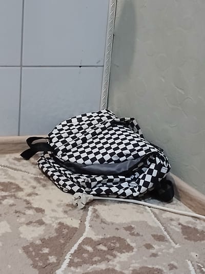 A checkerboard backpack is on the floor next to a wall and a patterned carpet. A white electrical cord with a plug is also on the carpet. The background consists of a tiled wall and a textured wall. There are no people present in the image. The location is an interior space, and there is no information to suggest it is located in the city of Korenovsk, Russia.