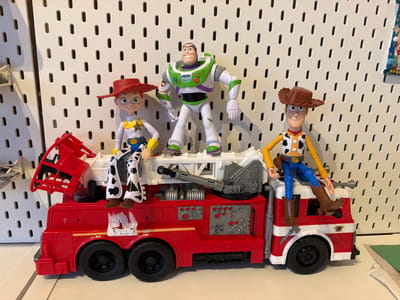 This media file features a collection of Toy Story action figures posed on a red toy fire truck. The figures include Jessie, Buzz Lightyear, and Woody. Jessie, on the left, wears her signature cow-print pants and red cowboy hat. Buzz Lightyear stands in the center, his iconic spacesuit detailed with green, purple, and white. Woody, on the right, is dressed in his cowboy attire, complete with a brown hat and vest.

The toy fire truck is bright red with white accents and features realistic details like wheels, ladders, and control panels. The action figures are positioned as if riding on or interacting with the truck. Jessie and Buzz are standing on the upper platform of the truck, while Woody is seated on the cab.

The background consists of a white pegboard with numerous small, evenly spaced holes, suggesting an indoor setting like a playroom or a store display. The lighting appears to be bright and even, consistent with an indoor environment. The overall scene is well-lit and the focus is clearly on the toys, creating a playful and colorful composition. No specific time of day or weather conditions are discernible, and the emotion conveyed is one of fun and play. The location is identified as Richmond, Canada.