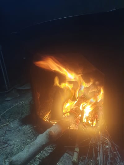 The image depicts an outdoor scene at night, likely in Восеабад, Tajikistan, featuring a bonfire. The fire burns brightly within a dark, possibly metal, enclosure, casting a warm glow on the surrounding area. Flames of orange and yellow dance upwards, illuminating logs and smaller branches within the fire pit. In the foreground, a thick log lies horizontally on the ground, with smaller sticks and debris scattered around it. The background is dark, with hints of vegetation or ground cover visible in the dim light. The overall impression is one of a rustic, nighttime setting centered around a crackling fire.