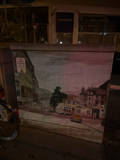 This is a nighttime photograph of a mural painted on the side of a building, likely an electrical box. The mural depicts an urban scene with buildings in the background, some with balconies and windows. A street with parked cars is also visible. The overall style of the painting is somewhat impressionistic, with soft colors and brushstrokes.

There is visible text on the mural, including "22 juillet 1966" written vertically on the left side, suggesting a historical date. Below this, a white sign with a yellow triangle and black exclamation mark, indicative of a warning, is also present. Text on this sign reads "COFFRET ELECTRICITE C 2538". Another sign with "VOLVO" in white text on a dark blue background is visible on the adjacent building to the left.

In the foreground, the wheel of a bicycle is visible, suggesting it might be parked nearby. The scene is lit by artificial light, indicating it is nighttime. The location is Geneva, Switzerland, as stated. The mood is calm and perhaps a bit nostalgic due to the historical date on the mural. The lighting and the mural's subject matter suggest an urban environment.