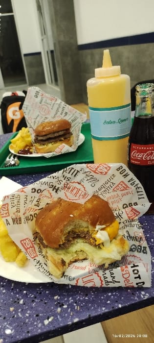 The image shows a tray with fast food items. There's a bottle of Coca-Cola and a squeeze bottle of "Salsa-Queso". Two burgers are visible, one mostly eaten and wrapped in paper printed with "HOT DOG" and "DELICIOUS" motifs. The other burger is smaller. French fries are served with both burgers. The background features a white and gray wall. A date and time stamp "16/02/2026 20:08" appear in the upper-right corner.