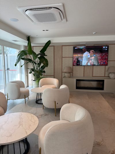 The image depicts an indoor lounge area with modern, neutral-toned furnishings, suggesting a commercial or hospitality setting in Les Duanes De La Mar, Spain. Prominently featured is a large, flat-screen television mounted on a wall, displaying a live sports broadcast, likely a soccer match, as indicated by the scoreboard and player uniforms. Two individuals are visible on the television screen. In the foreground and midground, several plush, cream-colored armchairs with gold-toned legs are arranged around marble-topped coffee tables. A large, tropical plant with broad green leaves stands in a white pot to the left of the television. The room is illuminated by natural light filtering through a large glass door on the left and by two decorative lamps placed on a credenza below the television. The credenza appears to house an electric fireplace. The flooring is a large-format, neutral-toned tile with a subtle marbled pattern. The ceiling features a recessed air conditioning unit and a textured, perforated surface. The overall ambiance is one of contemporary comfort and relaxation.