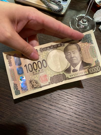 The image shows a close-up of a hand holding a Japanese 10,000 yen banknote. The banknote features a portrait of Yukichi Fukuzawa. The background is a dark wooden surface, likely a table. There is a glass of amber liquid and a decorative beaded bracelet visible in the upper right and upper left corners of the image, respectively. The lighting appears to be artificial, casting a shadow of the banknote onto the table. There is no discernible location in the city of Cherepovets, Russia.