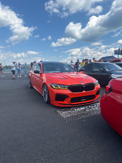 A bright orange BMW M5 is parked on an asphalt surface, with a group of people standing around it. The car is the main subject, facing slightly to the left. It has a black roof and a black grille, with yellow accents on the headlights. A license plate reading "CT 3686 TP" is visible.

In the background, a dark green car is parked behind the BMW. Further back, more people are visible, as well as tents and a fence. The sky is blue with scattered white clouds. The scene appears to be an outdoor car show or event. The location is stated to be Dospey, Bulgaria.