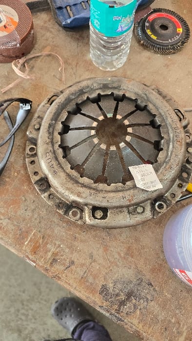 The image captures a close-up view of various objects arranged on a dirty and heavily scratched wooden workbench, likely in a mechanical workshop or garage setting in Gangapur, India.
The primary subject is a large, circular, metallic automotive clutch pressure plate assembly, positioned centrally. It appears used and covered in grime, with visible signs of wear. Engraved on its surface is the number "1300-0203," likely a part identification number, along with other faint, unreadable markings. A small, rectangular white tag is attached to the clutch assembly, featuring discernible text such as "SILICA," "OGI," and "KEEP AWAY FROM HEAT."
To the left of the clutch assembly, a clear plastic water bottle with a blue cap and label stands upright. Further left, a black and red circular flap disc or grinding wheel, marked "FLAP DISC," is visible. Below the water bottle, a smaller, brown abrasive disc, possibly a cutting wheel, is partially seen in its packaging, showing "MAX RPM" and other obscured text. A pair of black safety glasses or goggles rests on the bench near this disc.
In the upper right portion of the frame, a translucent purple container is partially visible. The lower right corner reveals a portion of a person's foot, clad in a black Crocs-style shoe.
The scene suggests an environment where maintenance or repair work is performed, with tools and automotive components laid out. No direct activity is taking place, but the context implies a practical, hands-on mechanical task.