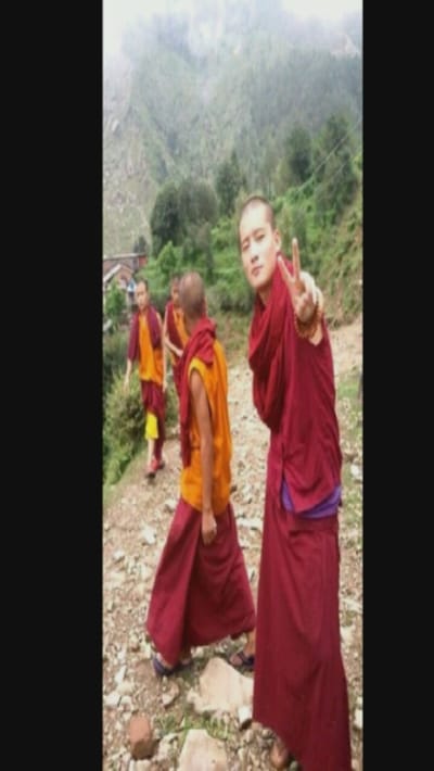 Four monks are walking along a path. Three are wearing robes with red and yellow. The fourth is in the foreground, wearing all red, and is making a peace sign. The background shows a mountain. The path appears to be rocky. The sky is overcast.